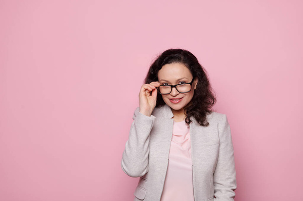 Beautiful dark-haired middle-aged woman in stylish elegant suit and spectacles, smiling looking at camera on isolated pink background. Eyesight. Health. Medicine. Ophthalmology and fashion concept - Photo, Image