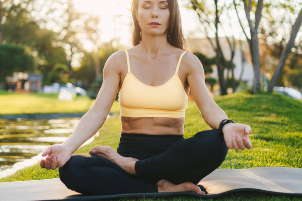 Retrato de una joven morena caucásica sentada en una esterilla de yoga meditando con los ojos cerrados en el parque. Postura de yoga, encontrar el equilibrio interior. Saludable - Foto, imagen