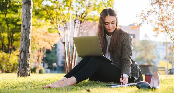Genç beyaz kadın, serbest çalışan iş kadını dizüstü bilgisayarda çalışıyor, açık havada parkta not defterine notlar yazan webinarları izliyor. Rahatlama yaşam tarzı kavramı - Fotoğraf, Görsel