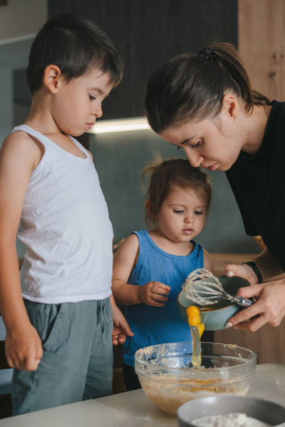 Cute caucasian kids and cheerful mother adding eggs in bowl while cooking in kitchen together. Family spending time together having fun. - Photo, Image