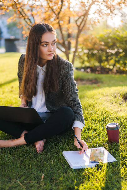 Młoda, atrakcyjna bizneswoman nosząca Jacke siedzącą na trawniku w parku w słoneczny dzień, pracująca nad swoją pracą dyplomową przy użyciu laptopa, wyglądająca na zajętą i - Zdjęcie, obraz