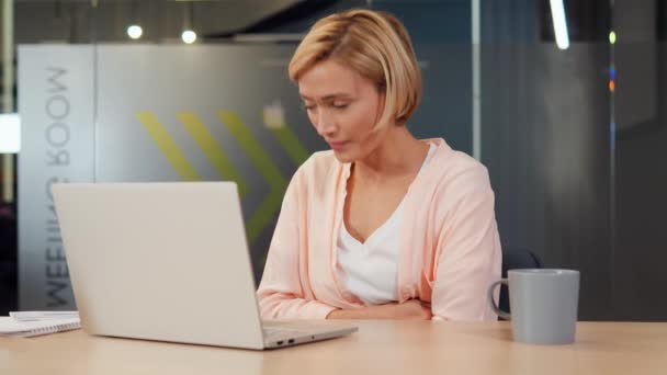 Portret zdeterminowanej blondynki pracującej nad laptopem pomimo bólu miesiączkowego w biurze. Dziewczyna prezentuje wyzwania, przed którymi stoją kobiety równoważące pracę i życie osobiste, nieznośny ból brzucha - Materiał filmowy, wideo