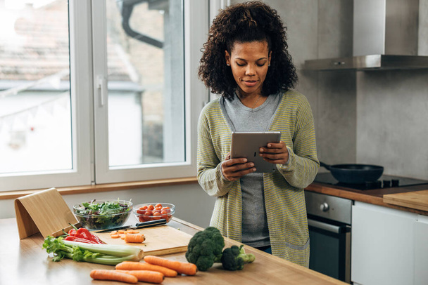 A woman is standing in the kitchen and reading a vegan recipe on tablet - Foto, afbeelding