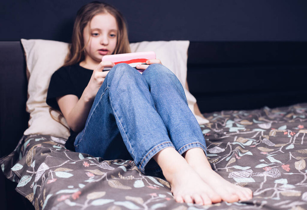 A teenage girl with bare feet is lying on the bed. Legs A child plays video games on the phone. Addiction to gadgets. - Foto, imagen