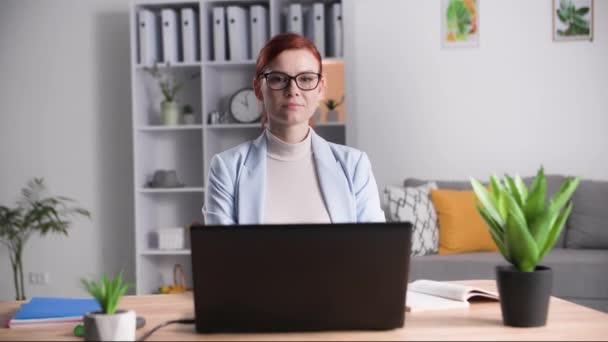 ritratto di una giovane donna con gli occhiali seduta davanti a un computer portatile nella stanza e che guarda la macchina fotografica, il concetto di home office o di lavoro a distanza - Filmati, video
