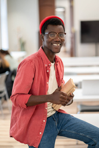 喜んでアフリカ系アメリカ人のヒップスター学生の男で眼鏡の図書館のテーブルの上に座っている本は親指を示すジェスチャー,推奨される効果的な大学教育のための手のような笑顔カメラを見て - 写真・画像