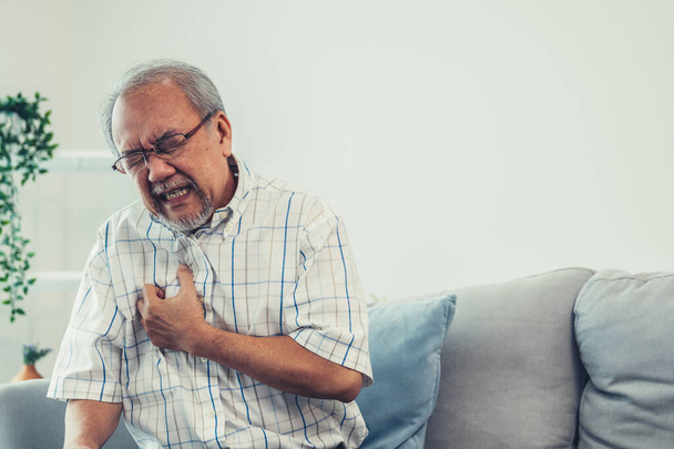 An agonizing senior man suffering from chest pain or heart attack alone in his living room. Serious health problem and feeling unwell concept for seniors. - Valokuva, kuva
