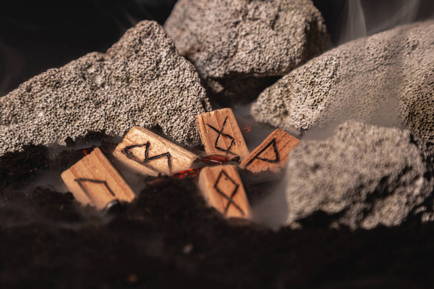 the wooden rune of Inguz among other runes, symbols of the earth, lie in the ground amid smoke and large stones. Mysticism and the rite of fruitful earth - Foto, afbeelding