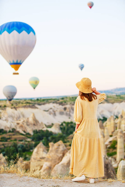 Young woman admires scenery of hot air balloons flying over Love valley with rock formations and fairy chimneys in Cappadocia Turkey - Photo, image