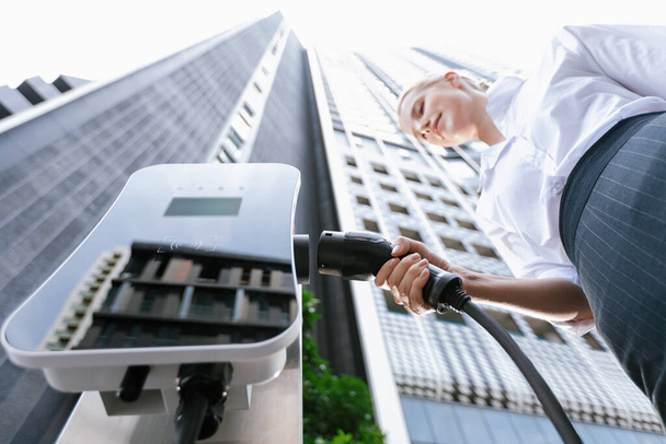 Fisheye view focus hand holding, inserting, plugging ev charger from public charging station for electric vehicle with blurred progressive businesswoman and residential city building in background. - Фото, зображення