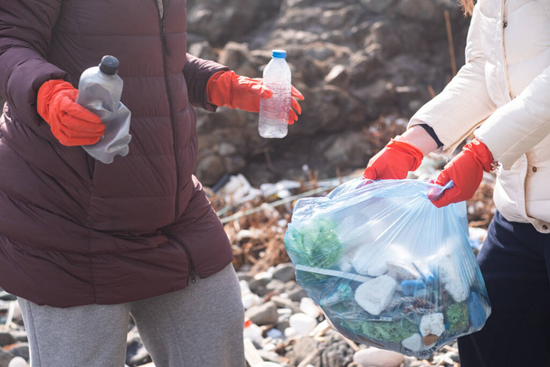 дві жінки збирають пластикові сміття в мішках на пляжі. Розлиті сміття навколо катастрофи забруднення. Порожні пластикові пляшки та відходи паперу
 - Фото, зображення