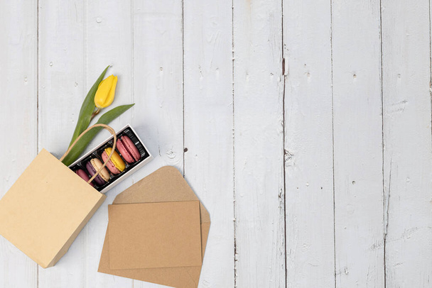 Una scatola di amaretti con un tulipano giallo in un sacchetto di carta e una busta vuota di kraf con un foglio si trovano a sinistra su un tavolo di legno bianco con spazio copia a destra, piatto lay close-up.. - Foto, immagini