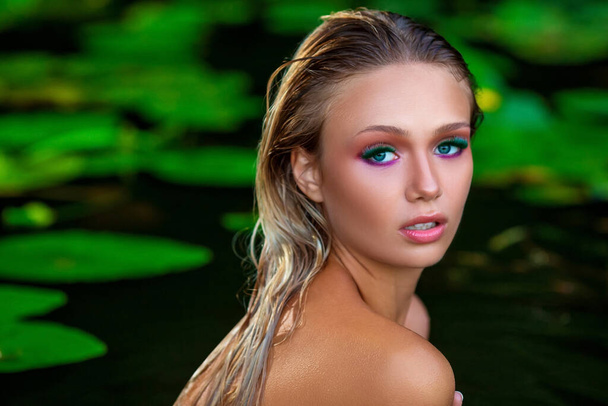 Close-up fashion beauty portrait of an attractive caucasian naked sexy blonde girl with creative greenish professional makeup shot in pond with green leaves half-drowned into the water - Φωτογραφία, εικόνα