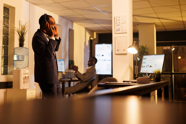 Der afroamerikanische Geschäftsmann trägt Kopfhörer, hört Musik und amüsiert sich im Startup-Büro. Geschäftsführender Manager genießt Arbeitspause, zeigt seine Tanzschritte nach Abschluss des Firmenprojekts - Foto, Bild