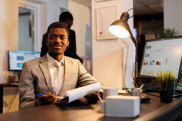 African american manager holding clipboard analyzing marketing statistics, working over hours at company strategy in startup office. Smiling business man looking at papers with investment graph - 写真・画像