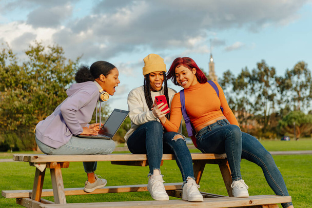 grupo multirracial de 3 amigas sentadas em um banco em um campus universitário usando um telefone celular e um laptop. colegas de classe se divertindo durante uma pausa - Foto, Imagem