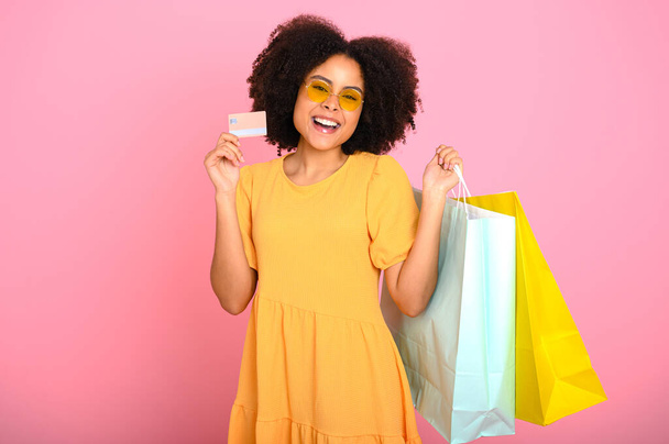 Pretty, happy, excited curly haired african american woman in an orange summer dress, holds shopping bags and credit banking card, looks at camera, smiles, stand on isolated pastel pink background - Foto, imagen