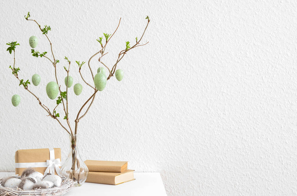 Vase avec branches d'arbre, oeufs de Pâques, coffret cadeau et livres sur la table près du mur de lumière - Photo, image