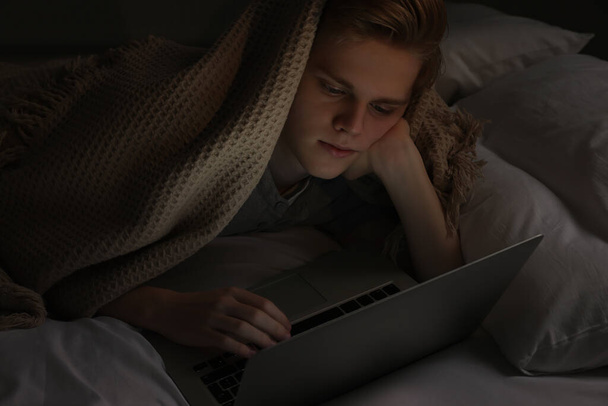 Teenage boy using laptop under blanket on bed at night. Internet addiction - Фото, изображение