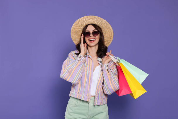 Όμορφη νεαρή γυναίκα με χάρτινες τσάντες για ψώνια σε μωβ φόντο - Φωτογραφία, εικόνα
