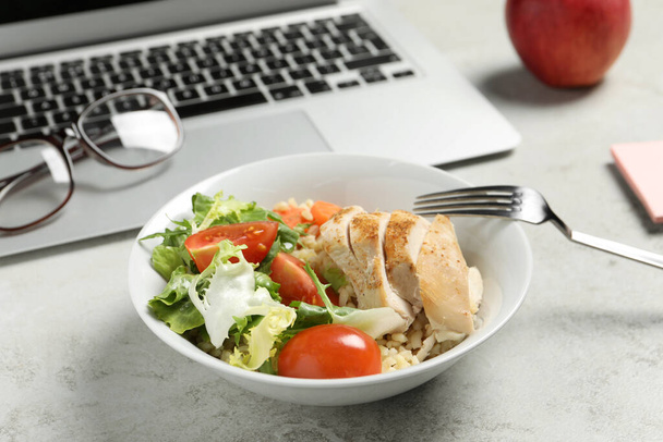 Bowl with tasty food, laptop, fork and glasses on light grey table, closeup. Business lunch - Фото, изображение