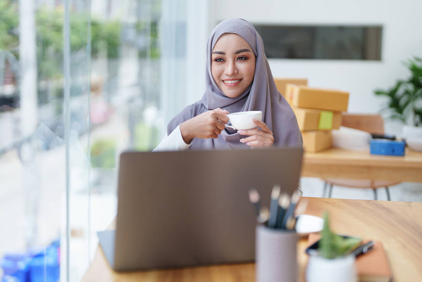 Μια όμορφη Μουσουλμάνα με χαμογελαστό πρόσωπο το πρωί πίνει καφέ και χρησιμοποιεί υπολογιστή.. - Φωτογραφία, εικόνα
