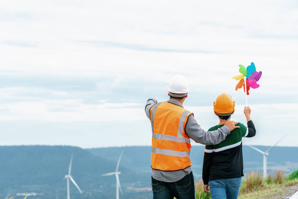 Engineer with his son holding windmill toy on a wind farm atop a hill or mountain. Progressive ideal for the future production of renewable, sustainable energy. Energy generated from wind turbine. - Foto, Imagem