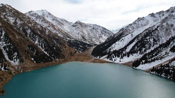 A lake in the mountains with turquoise blue water. Drone view of clear water, coniferous trees and snowy mountains. People walk along the shore, low bushes grow. Big Almaty lake. Kazakhstan - Zdjęcie, obraz