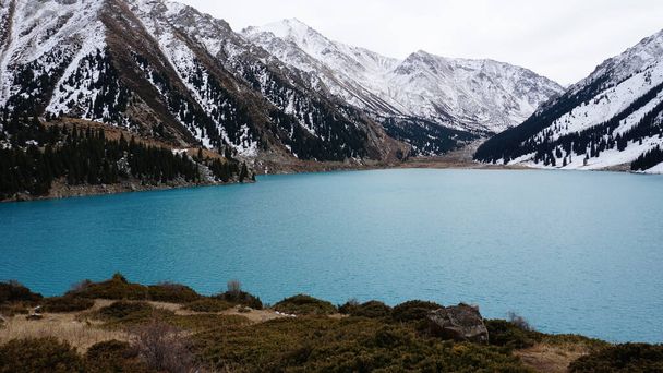 A mountain lake with blue water in winter. The suns rays are reflected from the water. Green forest and grass grow on the hills. There are stones lying. The high peaks are covered with snow. Almaty - Foto, imagen
