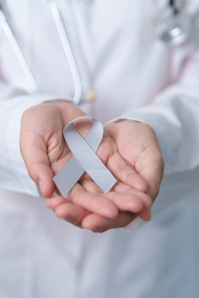 脳がん啓発5月、生活者をサポートするための灰色のリボンを持つ医師。医療と世界のがんの日のコンセプト - 写真・画像
