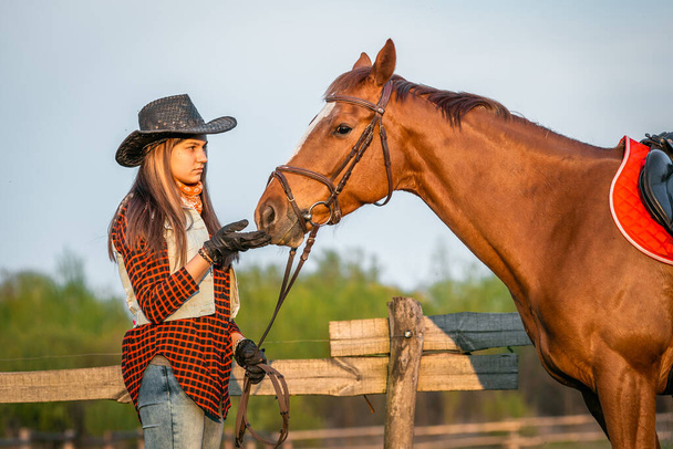 cowgirl in a hat standing near a horse in a field - Фото, зображення