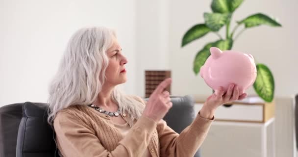 Starší žena s šedivými vlasy plísnění a mávání prstem na prasátko banky 4k film. Koncept malých důchodů - Záběry, video