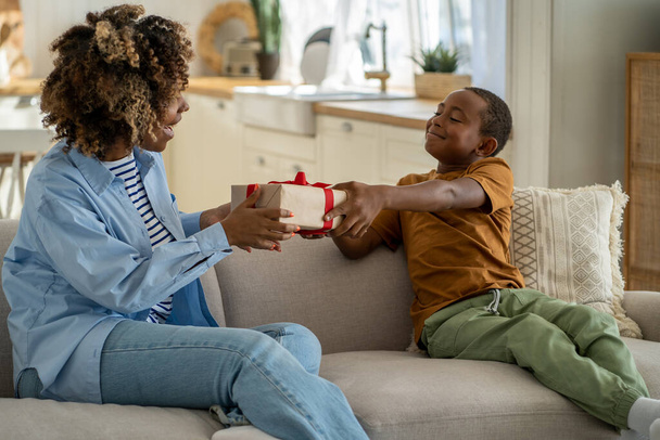 For the best mom ever. Proud happy African American boy son giving wrapped gift box to surprised excited mother, sitting together on couch at home. Child kid surprising mum on Mothers day - Φωτογραφία, εικόνα
