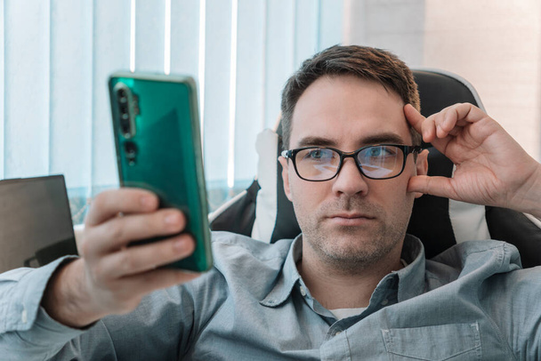 male use modern cellphone gadget, texting or messaging on wireless internet. Caucasian man in glasses sit at desk at home office look at smartphone screen consult client online. - Foto, afbeelding