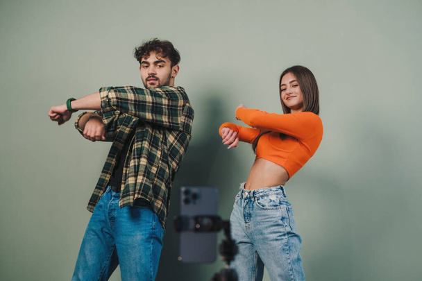 Positive young couple filming video blog for social network, using cell phone on tripod to broadcast their dance isolated on gray wall background. Content - Фото, изображение