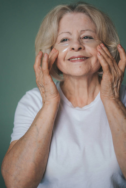 Sonriente vieja mujer caucásica con belleza natural aplicando parches bajo sus ojos. Cuidado de la piel de la cara en la vejez. Cara de belleza. Cuidado de la piel anti-envejecimiento. Belleza natural. - Foto, imagen