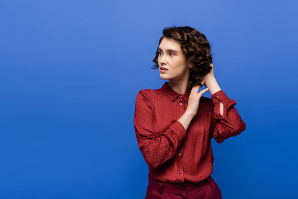 profesor de lenguaje reflexivo en blusa roja ajustando el pelo rizado y mirando hacia otro lado aislado en azul  - Foto, imagen