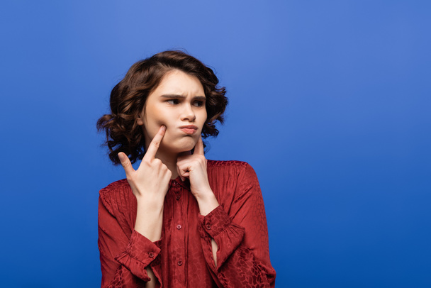 mujer joven disgustada señalando con los dedos las mejillas hinchadas mientras mira hacia otro lado aislado en azul  - Foto, Imagen