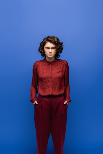 angry language teacher in maroon color pants standing with hands in pockets isolated on blue  - Foto, Imagem