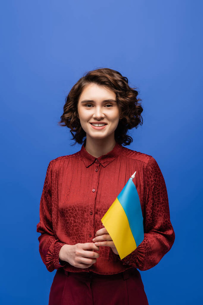 happy language teacher holding flag of Ukraine and smiling isolated on blue  - Fotó, kép