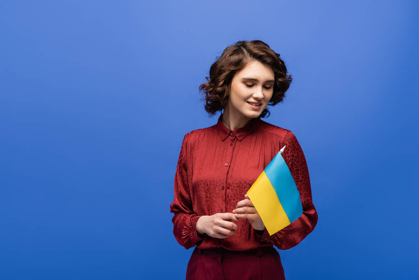 χαρούμενη γλώσσα δάσκαλος κρατώντας σημαία της Ουκρανίας και χαμογελώντας απομονωμένη στο μπλε  - Φωτογραφία, εικόνα