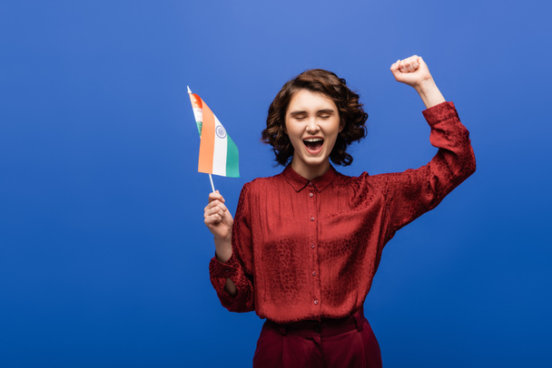 mujer excitada con el pelo corto rizado sosteniendo la bandera de la India aislado en azul  - Foto, Imagen