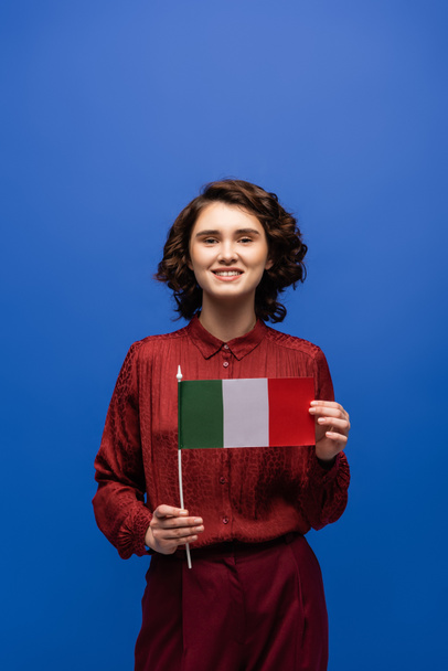 feliz profesor de idiomas sonriendo mientras sostiene la bandera de Italia y mirando a la cámara aislada en azul  - Foto, imagen