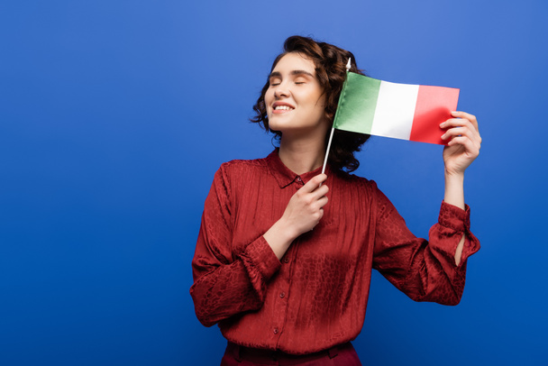 приємна вчителька мови посміхається, коли тримає прапор Італії ізольованим на синьому  - Фото, зображення