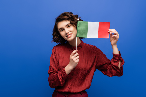 šťastná žena se usmívá, zatímco drží vlajku Itálie a dívá se na kameru izolované na modré  - Fotografie, Obrázek