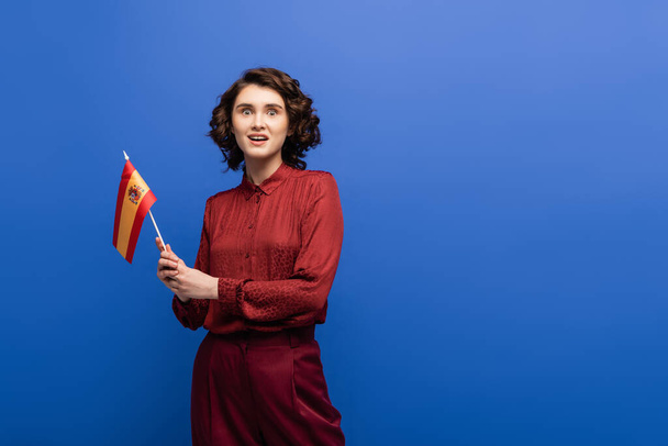 verblüffter Sprachlehrer blickt in die Kamera, während er die Flagge Spaniens isoliert auf Blau hält  - Foto, Bild