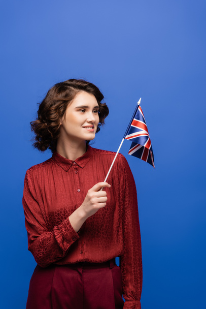 feliz profesor de idiomas sonriendo mientras mira la bandera de Reino Unido aislada en azul  - Foto, Imagen