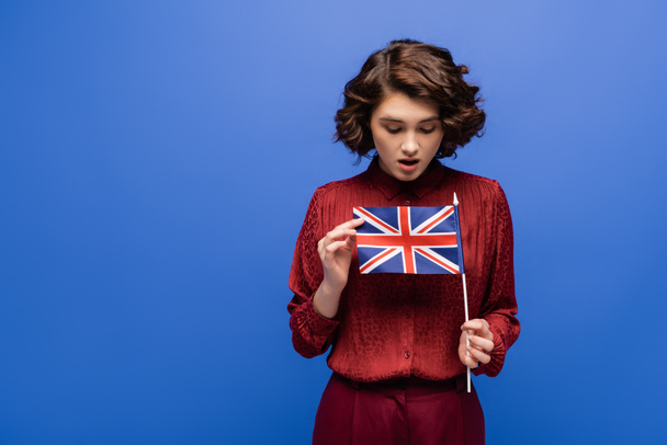σοκαρισμένος μαθητής με σγουρά μαλλιά κοιτάζοντας σημαία του Ηνωμένου Βασιλείου απομονωμένο σε μπλε  - Φωτογραφία, εικόνα