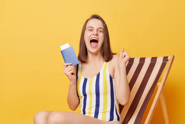 Horizontal shot of amazed excited overjoyed young woman wearing colorful swimsuit, holding tickets and passport in hands, yelling with happiness, sitting on wooden chair isolated on yellow background - Photo, Image