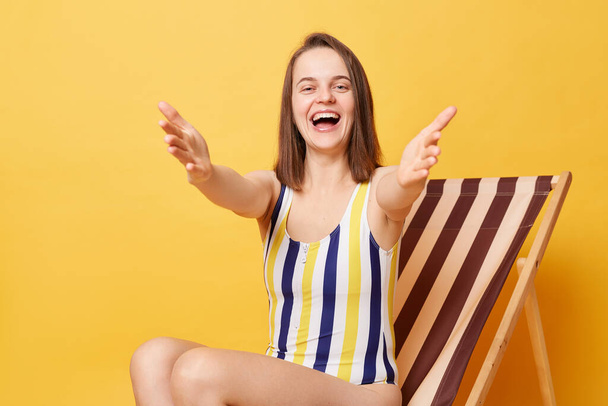 Portrait of friendly happy young woman wearing striped one-piece swimsuit sitting on wooden chair isolated on yellow background, inviting you to resort, saying come into my arms - Фото, зображення